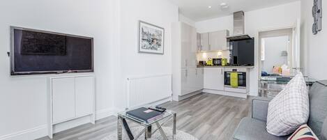Una sala de estar moderna con un sofá, una mesa de centro de vidrio, un televisor de pantalla plana y una zona de cocina al fondo.