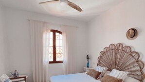 Une chambre à coucher comprenant un lit, un ventilateur de plafond, une fenêtre avec des rideaux, une table de chevet et un chapeau accroché au mur.