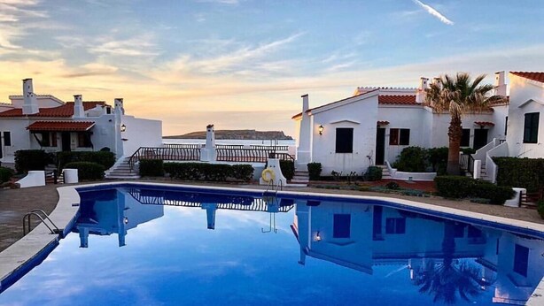 Une piscine entourée de bâtiments blancs aux toits recouverts de tuiles rouges, un ciel dégagé et un coucher de soleil.