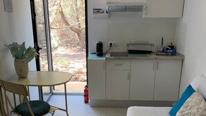 Una habitación compacta con cama, una mesita con una silla, kitchenette y una puerta corrediza de vidrio que da a un espacio al aire libre.
