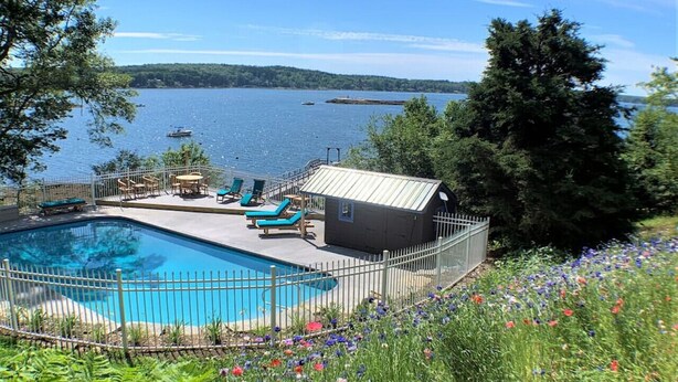 A swimming pool with lounge chairs and a small shed, surrounded by a fence and a flower bed, overlooking a lake. - Waterfront Cabin with Inground Pool, Dock, Tennis (Boothbay Harbor)