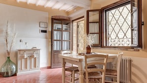 Un coin repas avec une table et des chaises en bois, un vase contenant des plantes séchées, un meuble avec des serviettes suspendues et une fenêtre ornée de motifs en fer forgé.