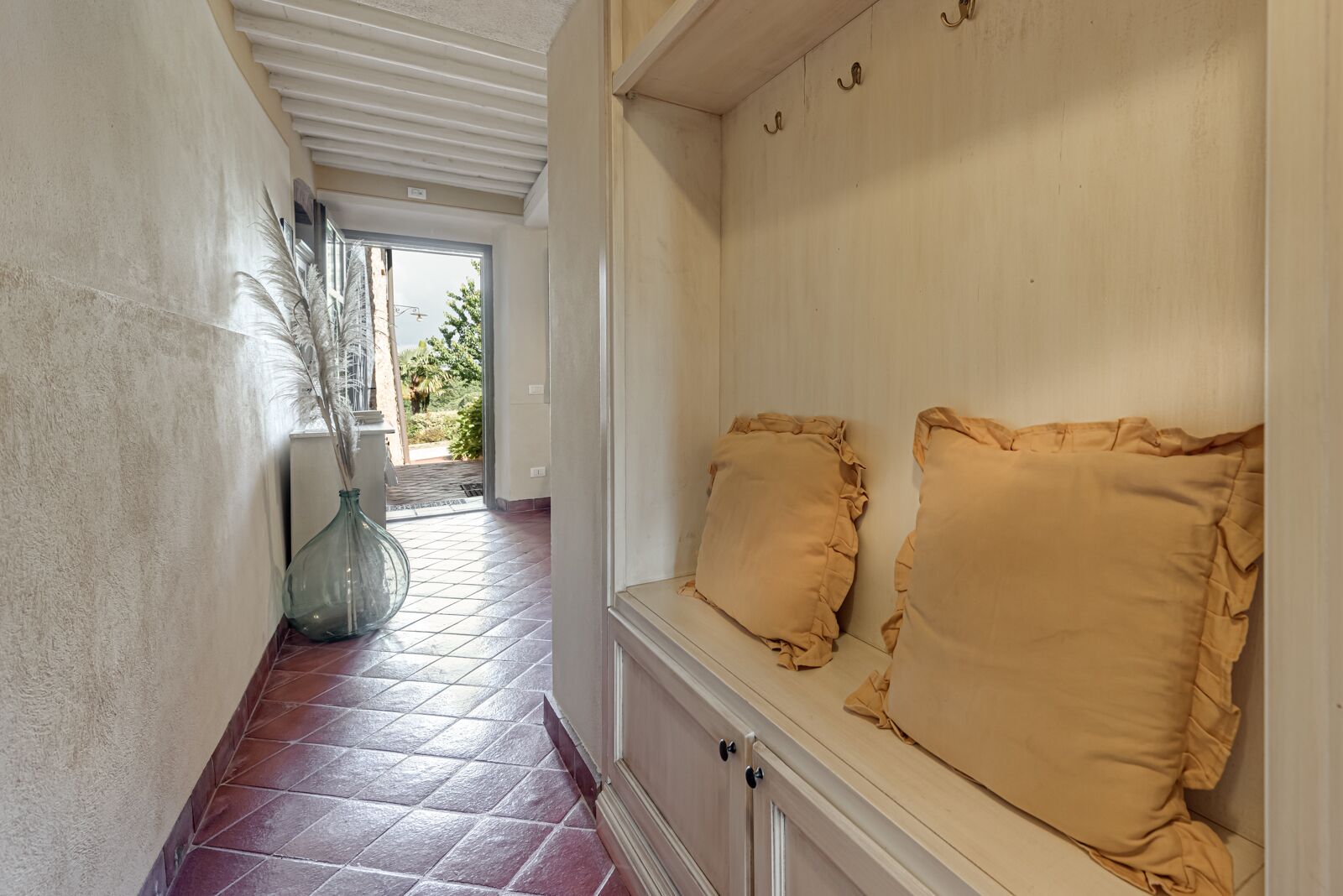 A hallway with a bench, a vase with dried plants, and a view of the outdoors.