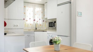 A compact kitchen with white appliances, a wooden table, and a decorative window treatment. - Orquidea Apartment - Historical Center (Málaga)