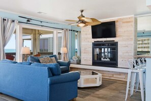 A living room with a blue sofa, a fireplace, a ceiling fan, and a TV mounted on the wall. - 5736-Sea Sunrise, | Oceanfront | Elevator Access | Diamond Shoals | Nags Head | Sleeps 6 (Nags Head)