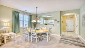 A dining area with a round table, six chairs, a chandelier, and a window with curtains. - 1024 River Crossing - Barefoot Resort Rentals (North Myrtle Beach)