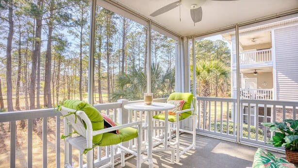 A covered outdoor area with two chairs and a table, surrounded by a white railing, overlooking a forested area. - 1024 River Crossing - Barefoot Resort Rentals (North Myrtle Beach)
