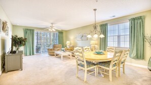 A spacious living room with a dining area, large windows with curtains, a ceiling fan, and a view of the outdoors. - 1024 River Crossing - Barefoot Resort Rentals (North Myrtle Beach)