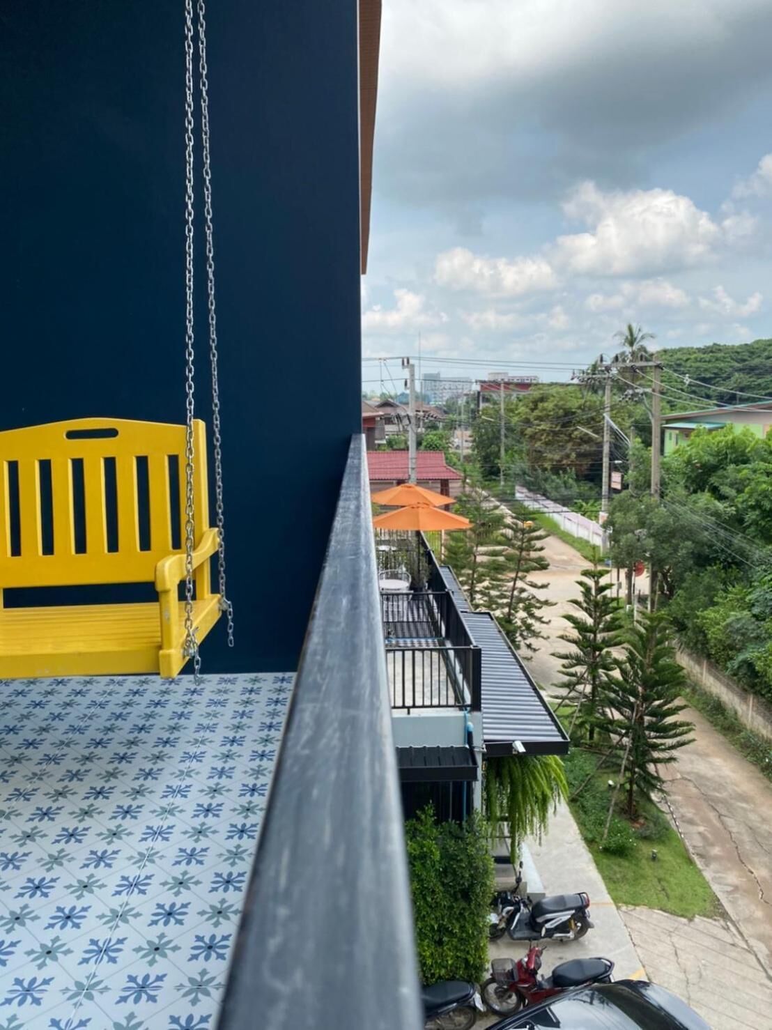 Un balcón con una silla amarilla, un piso con diseño y vistas a una calle con zonas verdes y edificios.