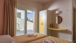 Een hotelkamer met twee bedden, een schuifdeur naar buiten, een geel schap, een ronde spiegel en een kleine vaas met bloemen.