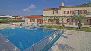 Une piscine entourée de chaises longues, un bâtiment en pierre avec des toits couverts de tuiles rouges et un ciel bleu azur.