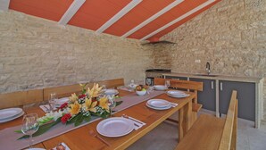 Un espace repas avec une longue table en bois dressée pour le repas, des murs en pierre et un coin cuisine équipé d’un évier et d’une cuisinière.