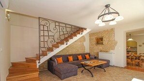 Un escalier avec des marches en bois et des rampes en métal, un canapé d’angle gris avec des coussins orange et une table basse en bois dans un salon aux murs en pierre, avec une cuisine en arrière-plan.