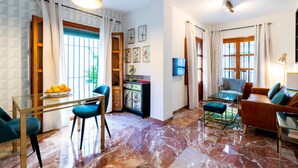 Amplia sala de estar con una mesa de centro de vidrio, sillas azules, un sofá de cuero y una mesa de comedor con sillas. Hay ventanas grandes con cortinas, un candelabro y obras de arte en las paredes.