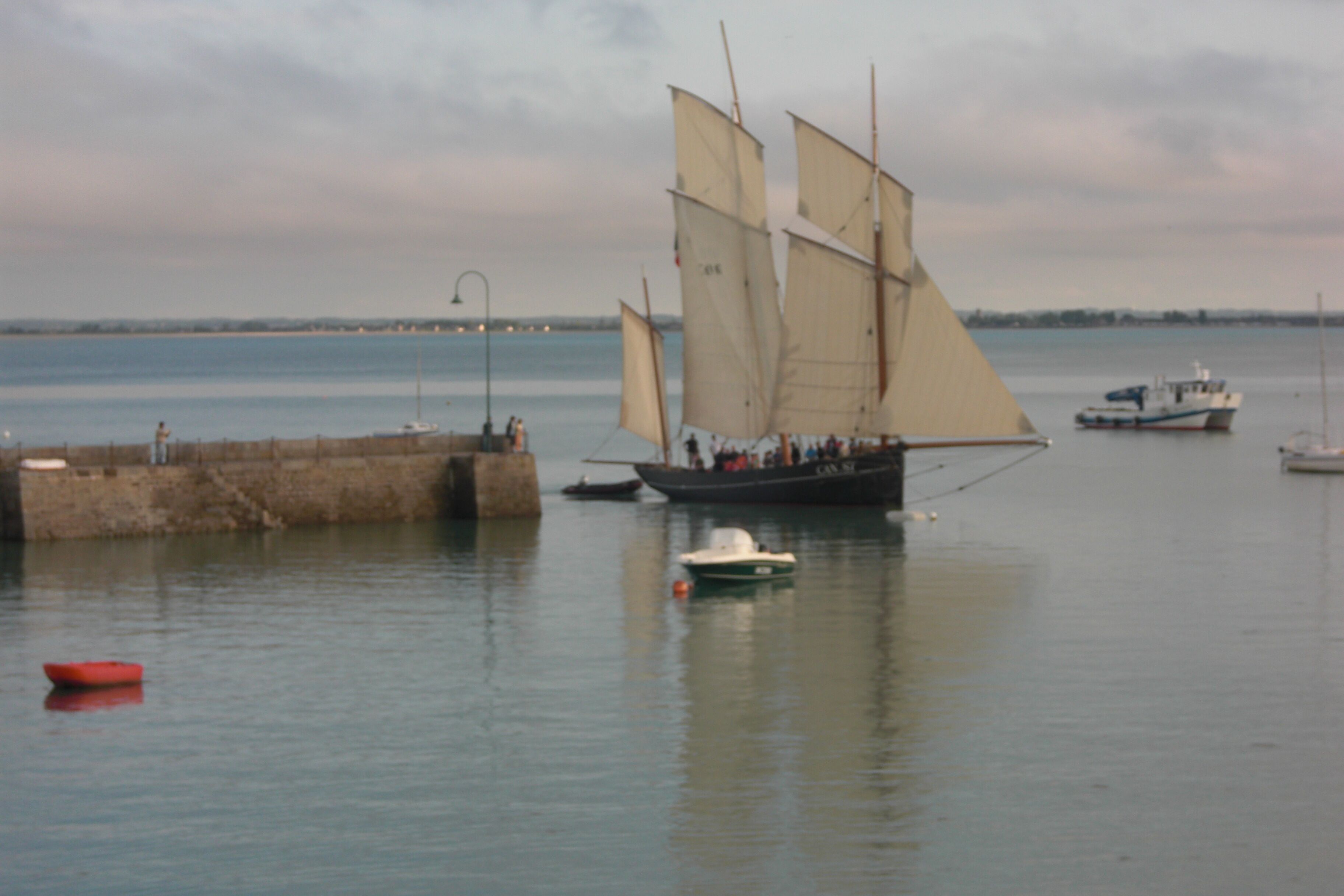 A harbor with several sailboats, including a prominent sailboat with multiple sails, anchored near a stone pier.