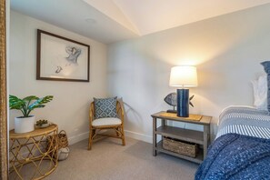 A bedroom with a bed, a chair, a small table with a lamp, a framed picture on the wall, and a potted plant.