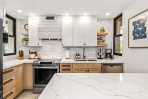 A modern kitchen with white cabinetry, a marble countertop, stainless steel appliances, and a window providing natural light. - Wailea Elua #102 Beautifully Remodeled Ocean View short walk to beach, shops! (Wailea)