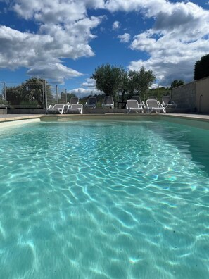 Una piscina limpia con sillas de descanso blancas alrededor, una zona vallada y árboles al fondo.