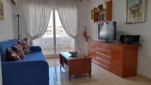 A living room with a blue sofa, a wooden cabinet, a television, and a balcony with white curtains. - Perfect views and location (Oropesa)