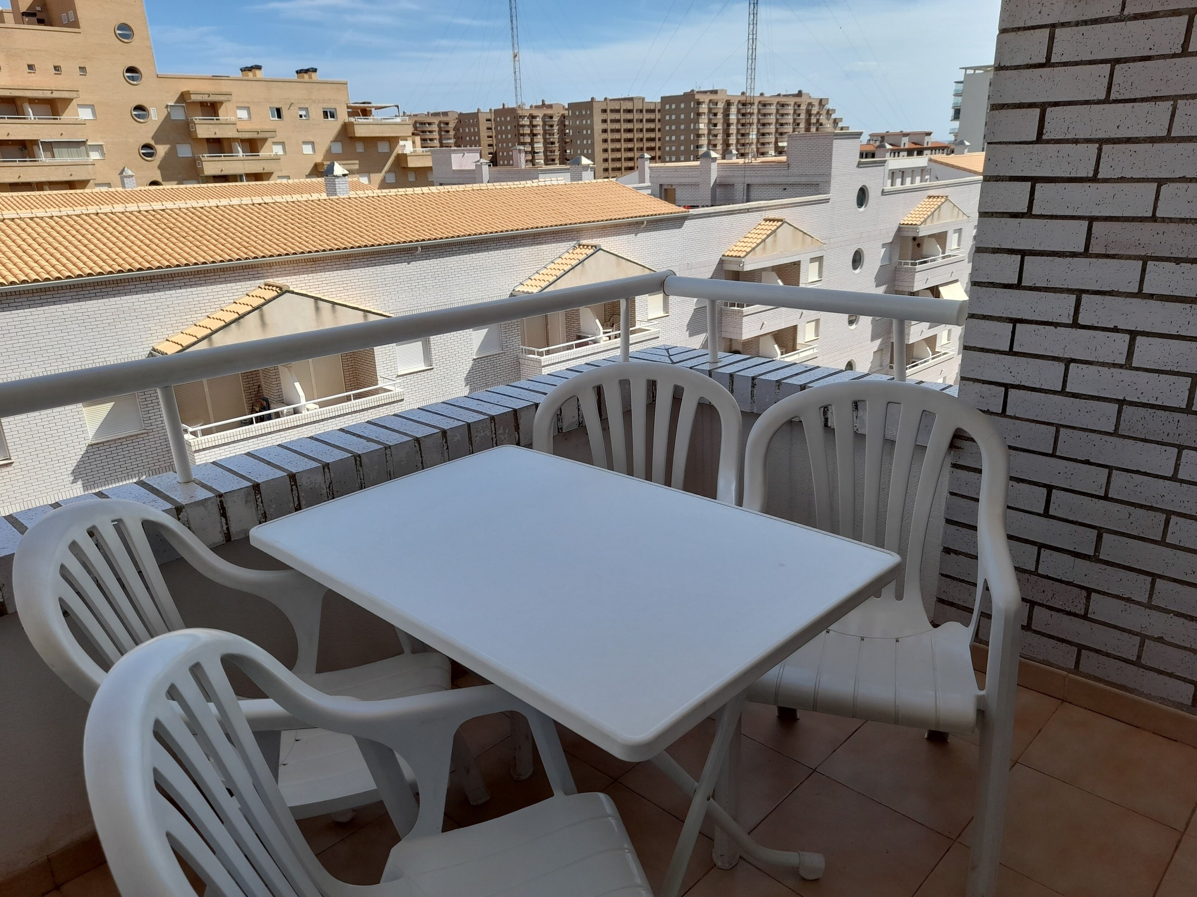 Un balcon avec une table et des chaises blanches, donnant sur un paysage urbain composé d’immeubles et d’un ciel dégagé.