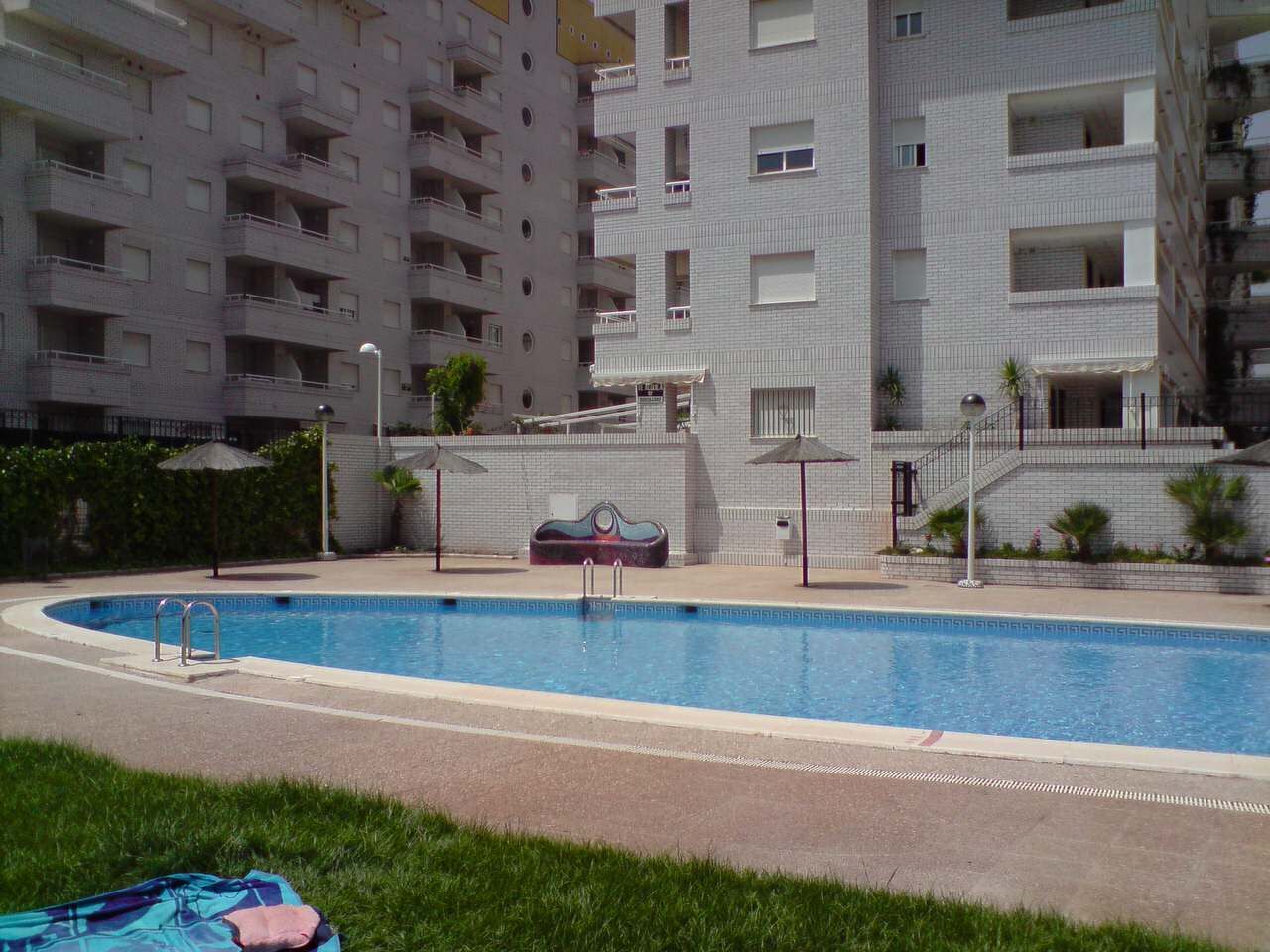 A swimming pool with a tiled deck, surrounded by a building with multiple balconies and a covered staircase.