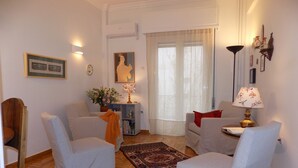Ein Wohnzimmer mit zwei Sesseln, einem Sofa, einem kleinen Tisch mit Lampe, einem Bücherregal und einem großen Fenster mit durchsichtigen Vorhängen.
