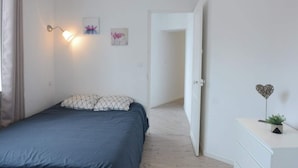 A bedroom with a bed, a nightstand, a wall-mounted lamp, and a door leading to another room. - Perle Marine - Dieppe (Dieppe)