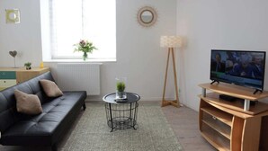 A modern living room with a black leather sofa, a small round coffee table, a TV on a wooden stand, and a large window with a view of greenery outside. - Perle Marine - Dieppe (Dieppe)