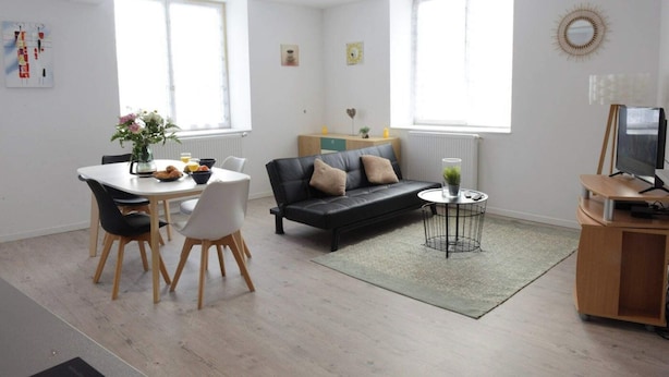 A modern living room with a black leather sofa, a dining table with chairs, a TV stand, and a large window providing natural light. - Perle Marine - Dieppe (Dieppe)