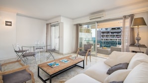 Una sala de estar moderna con una mesa de vidrio para comer, un sofá y una mesa de centro con revistas. Posee una puerta corrediza de vidrio que da a un balcón con mobiliario exterior.