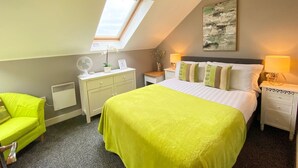 Une chambre avec une lucarne, un lit recouvert d’une couverture verte, une commode blanche, une table de chevet avec une lampe et un tableau accroché au mur.