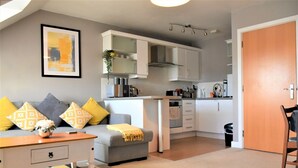 Un espace de vie compact comprenant un coin cuisine, un canapé gris agrémenté de coussins jaunes, une table basse en bois et un tableau encadré accroché au mur.