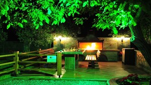 Une terrasse extérieure dotée d’une cheminée en pierre, de sièges en bois et d’un jardin illuminé de lumière verte.