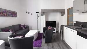 Un salon moderne avec un canapé gris, une table basse blanche et un fauteuil noir. Un tapis violet recouvre le sol, et un tableau est accroché au mur.