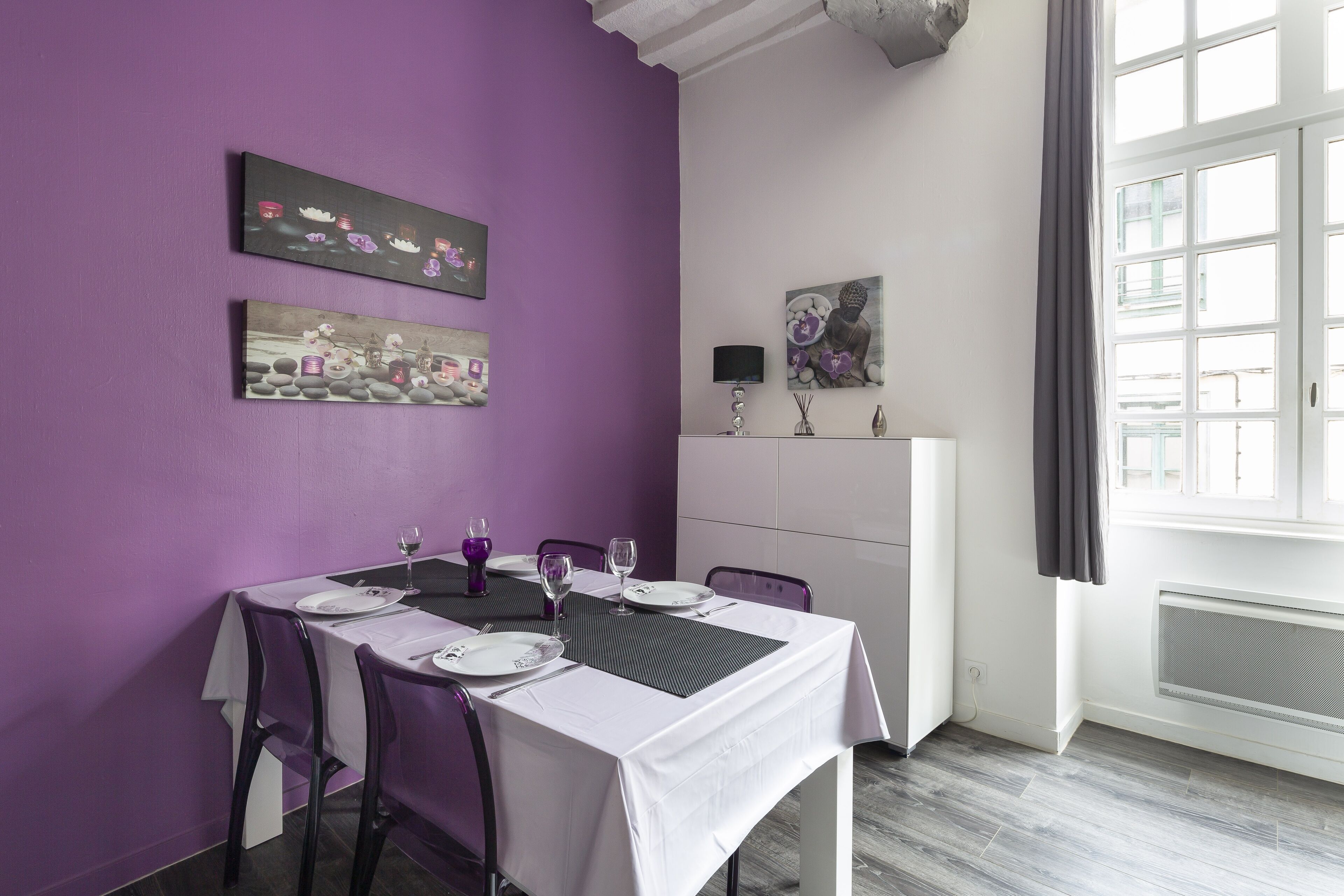 A dining area with a purple wall, white tablecloth, and modern chairs. There are paintings on the wall and a window with curtains.