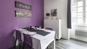 Un coin repas avec un mur violet, une nappe blanche et des chaises modernes. Des tableaux ornent le mur, et une fenêtre est agrémentée de rideaux.