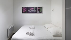 Un lit bien fait, avec des serviettes pliées, un mur blanc et un tableau encadré au-dessus.