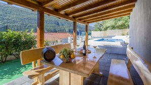 Uma pérgula de madeira ao ar livre com uma mesa posta para refeições, com vista para uma piscina e montanhas.