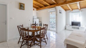 Uma área de refeições com mesa e cadeiras de madeira, uma cozinha ao fundo e um quarto com televisão e janela.