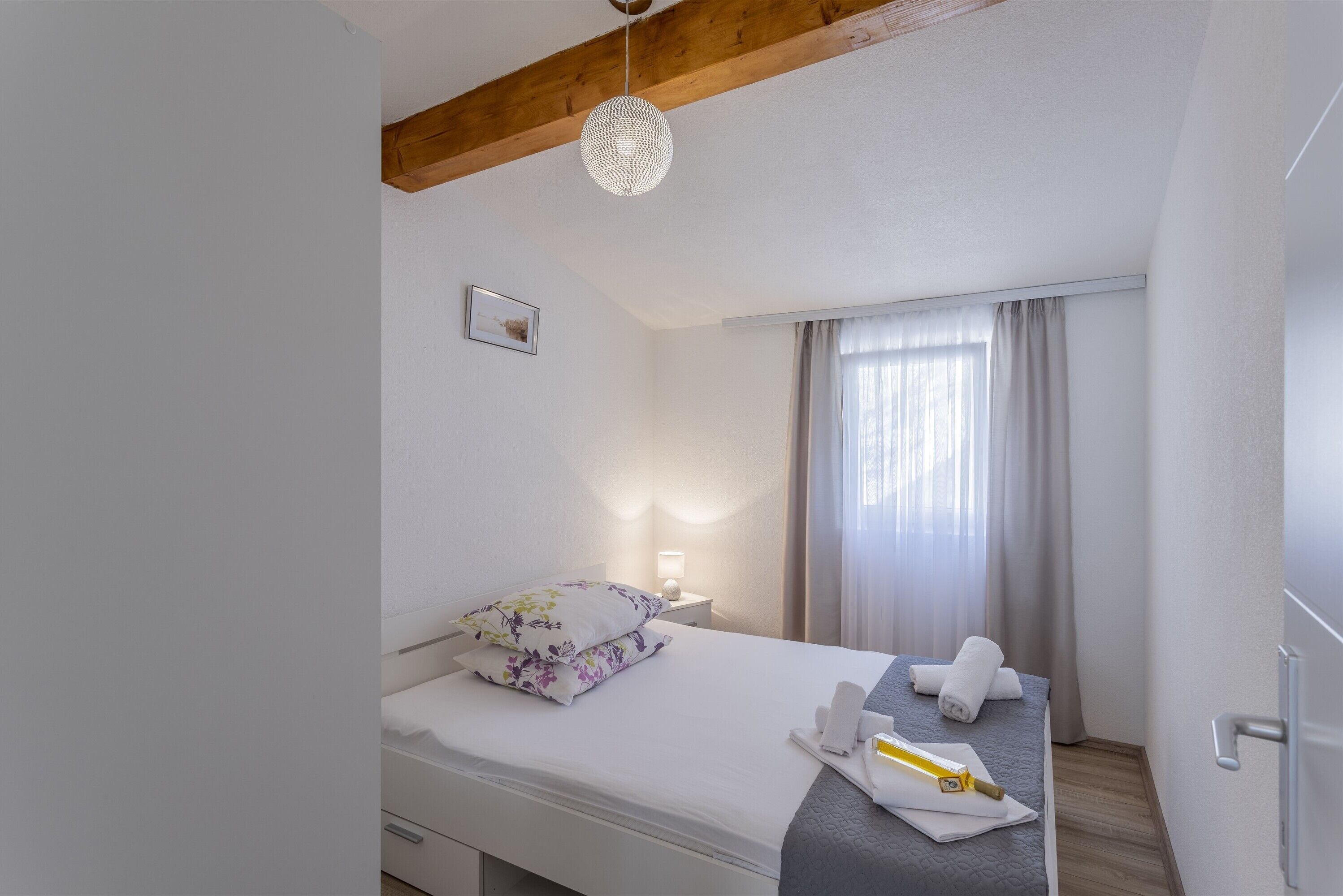 A hotel room with a bed, a bedside table, a lamp, a window with curtains, and a wooden ceiling beam.