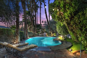 A swimming pool with lounge chairs, surrounded by tall trees and a sunset sky.