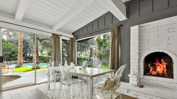 A modern dining area with a glass table and chairs, a fireplace, and a view of a swimming pool and garden. - Luxury Palm Desert Mid-Century Home Private Pool Golf Course (Palm Desert)