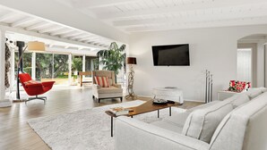 A spacious living room with a white sofa, red chair, and a coffee table. A TV is mounted on the wall, and there is a large window providing natural light. - Luxury Palm Desert Mid-Century Home Private Pool Golf Course (Palm Desert)