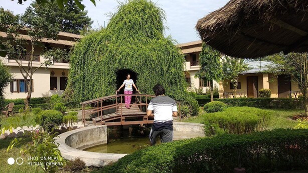 A courtyard with a wooden bridge, a pond, and buildings with thatched roofs. - Bardia Tiger Resort (Dhadhawar)