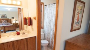Kylpyhuoneessa on puinen allaskaappi, wc-istuin, suihkuverho ja seinällä oleva kehystetty kuva.