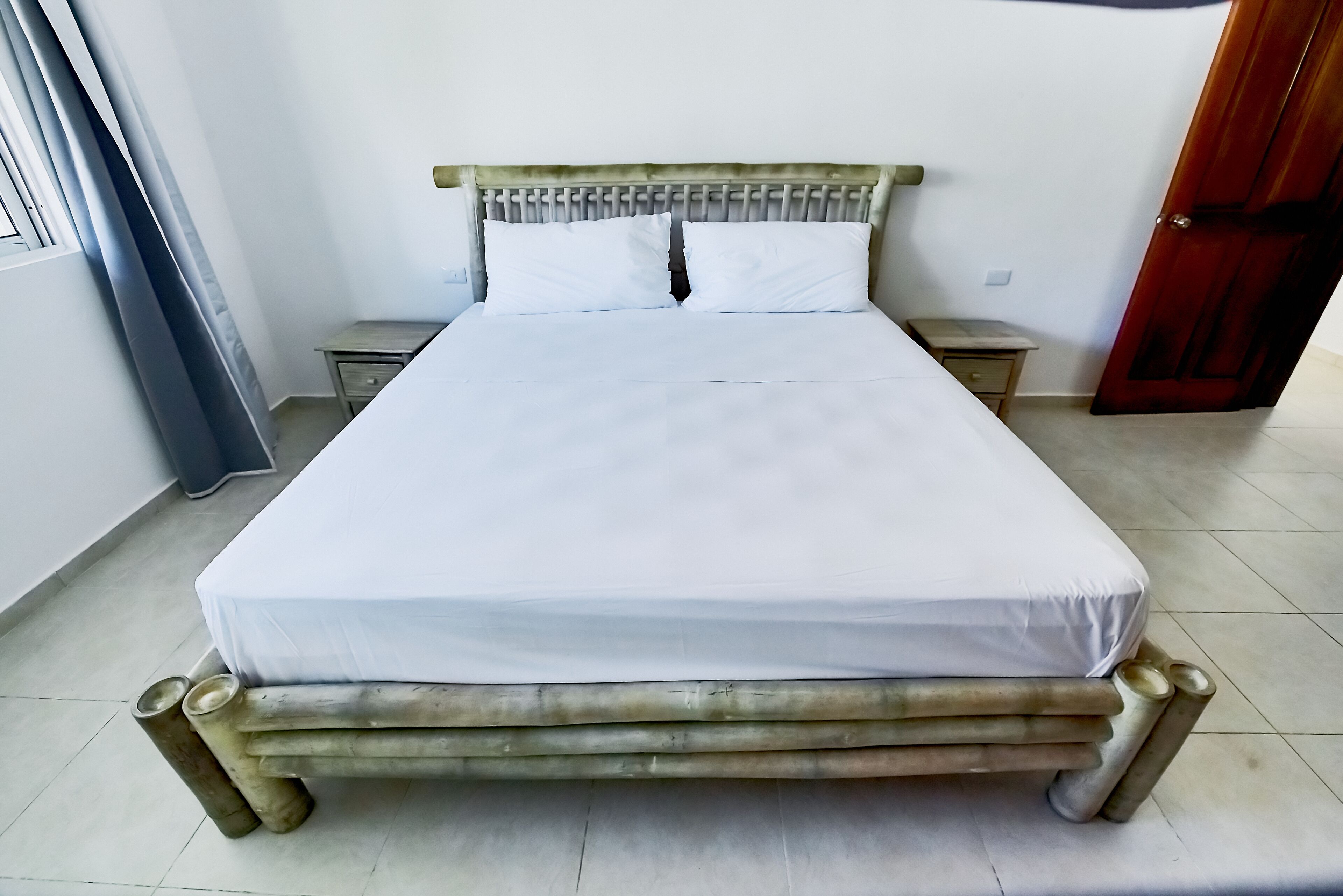 A bed with a wooden frame and white bedding in a room with a tiled floor and a wooden door.