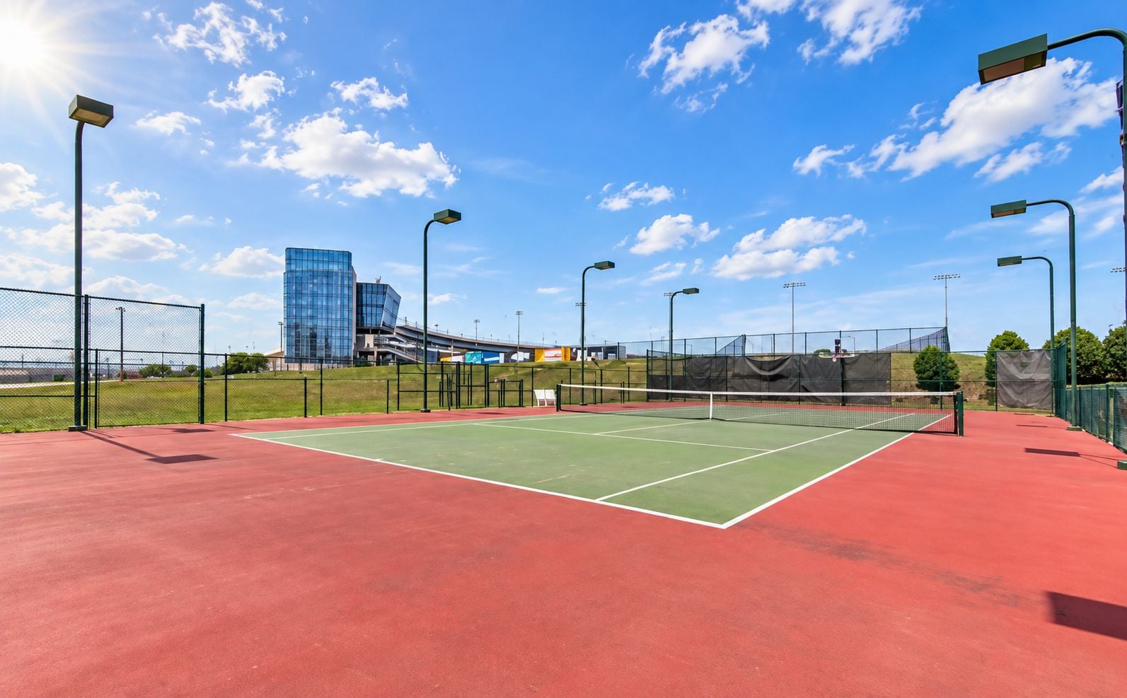 Un court de tennis extérieur doté d’une surface rouge, entouré d’une clôture verte, avec un bâtiment moderne en arrière-plan.