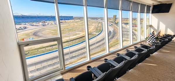 Una habitación con ventanas amplias que dan a una pista de carreras, filas de sillas negras y un cartel de NASCAR.