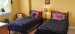 Un dormitorio con dos camas individuales, una mesita de noche, una lámpara, una silla, una ventana con persianas y un cuadro en la pared.