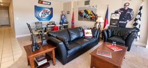 Ein Wohnzimmer mit einem schwarzen Ledersofa, einem Couchtisch und einem Esstisch mit Stühlen. An den Wänden hängen Banner von NASCAR und Miller Lite.